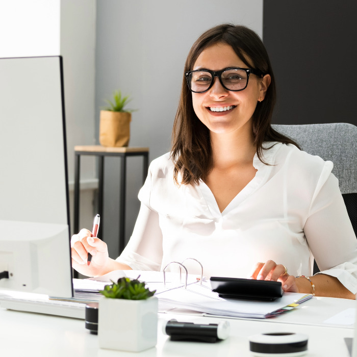 A woman with dark, thick rimmed glasses smiling in front a large computer monitor